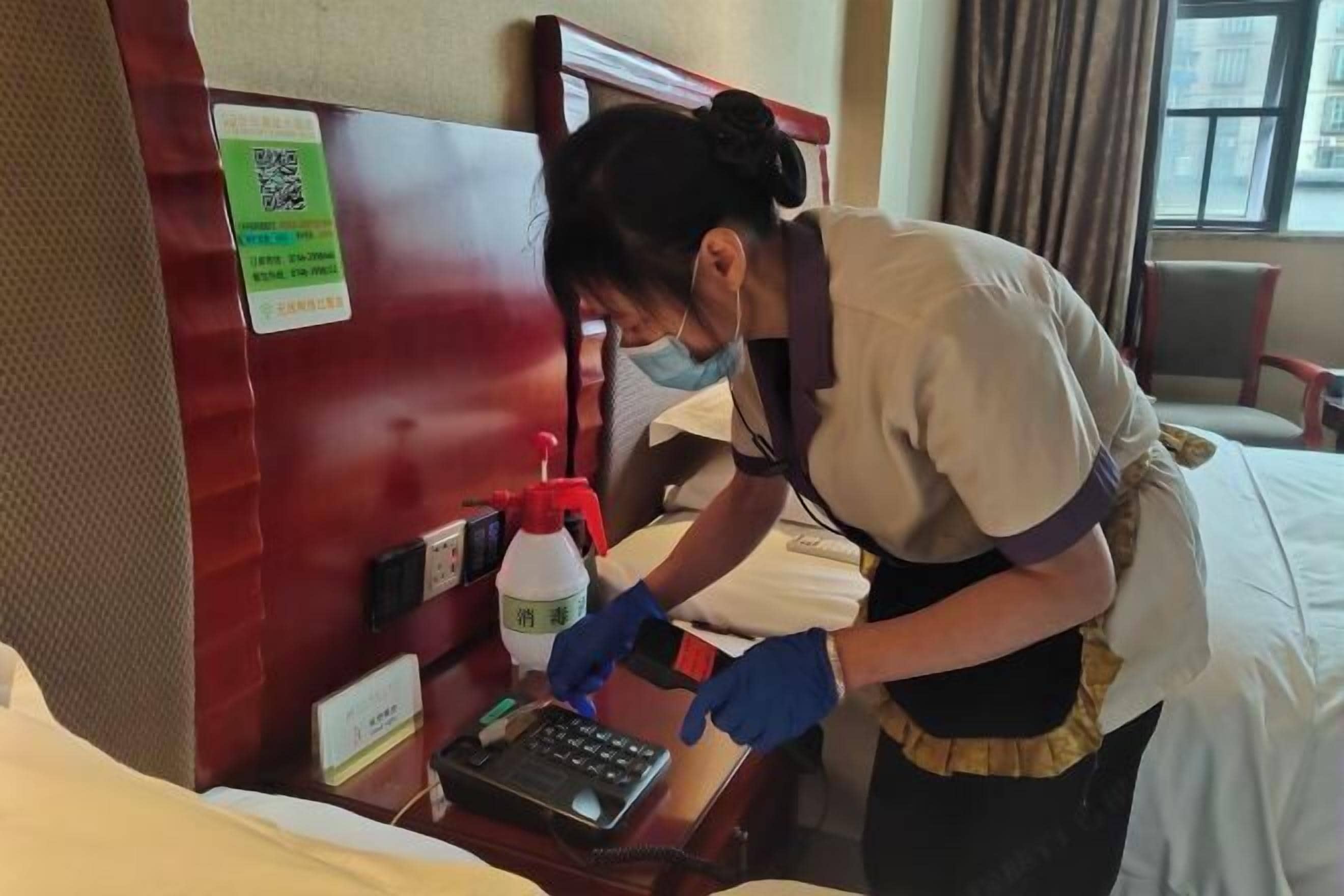 A hotel room with a bed, a desk, and a nightstand. There is a QR code on the wall, a bottle of hand sanitizer, and a phone on the desk.