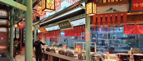 A food stall with a variety of dishes displayed in front of a glass enclosure, a person serving, and traditional decorations.