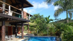 A pool area with a wooden deck, outdoor seating, and lush tropical vegetation.