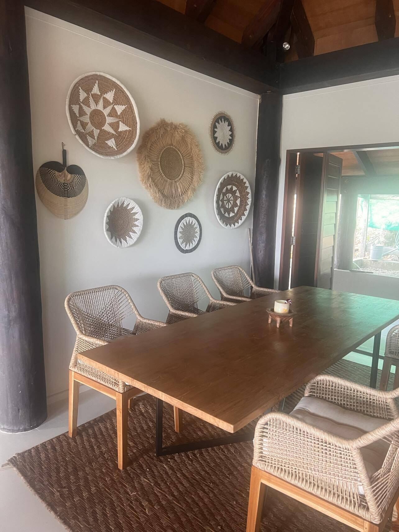 A dining area with a long wooden table surrounded by wicker chairs, a woven rug, and decorative plates on the wall.