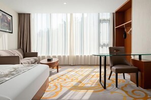 Camera d'albergo con un letto, una poltrona, un tavolino in vetro, una sedia e vista sulla città attraverso ampie finestre.
