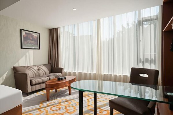 Habitación de hotel con una mesa de vidrio, un sofá, una silla y un ventanal con cortinas.