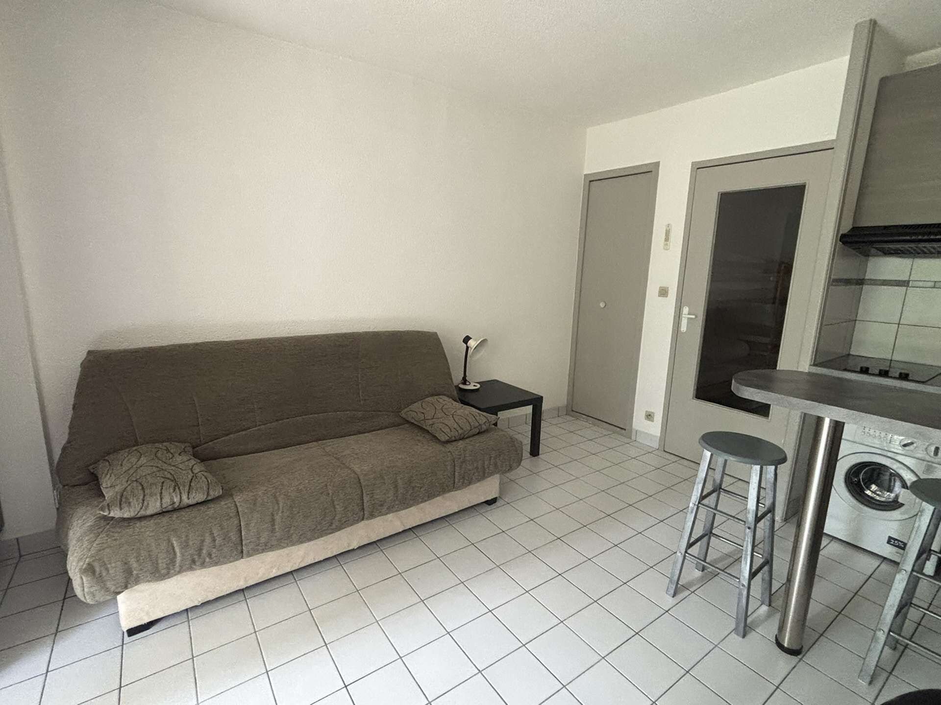 Ett kompakt bostadsutrymme med en soffa, ett litet bord, en tvättmaskin och ett litet kök.