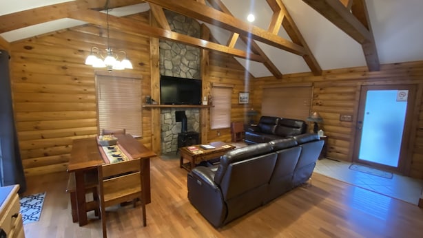 A log cabin interior with a wooden dining table, a leather sofa, a stone fireplace, and a television mounted on the wall. - Bear Timbers log cabin on the Shenandoah River (Rileyville)