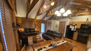A cozy living room with a wooden ceiling, a black leather sofa, a wooden dining table with a placemat, and a kitchen area with wooden cabinets and appliances. - Bear Timbers log cabin on the Shenandoah River (Rileyville)