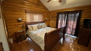 A log cabin bedroom with a wooden bed, a ceiling fan, a window with blinds, and a television on a wooden cabinet. - Bear Timbers log cabin on the Shenandoah River (Rileyville)