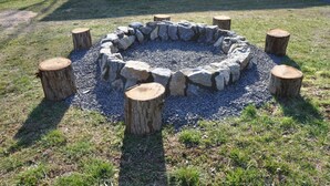 A circular stone fire pit surrounded by wooden stumps, set on a grassy area with a building in the background. - Bear Timbers log cabin on the Shenandoah River (Rileyville)