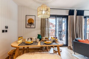 Uma sala de jantar com mesa de madeira posta para o café da manhã, luminária pendente e vista para o exterior através de grandes janelas.
