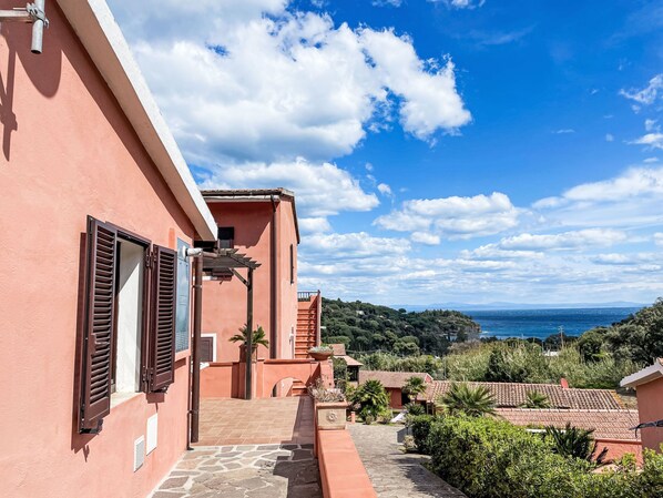 Un bâtiment rose avec des volets, un chemin de pierre et une vue sur la mer.