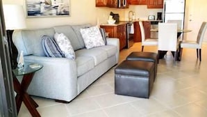 Un salon avec un canapé gris, une petite table et un pouf noir. Il y a également un coin cuisine avec des meubles en bois et un réfrigérateur.
