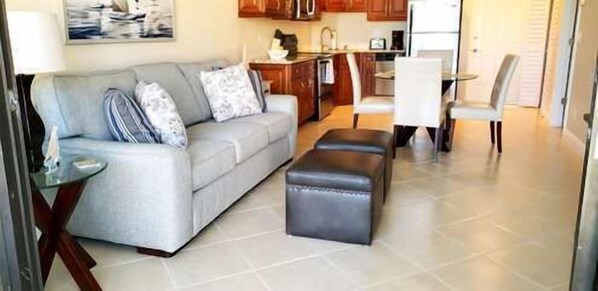 A living room with a grey sofa, a small table, and a black ottoman. There is a kitchen area with wooden cabinets and a refrigerator. - GV 5325 Golf Course View Condo-Paradise Vacation Rental in Ocean Village (Fort Pierce)