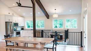 Een ruime keuken met moderne apparatuur, een eethoek met een houten tafel en stoelen, en grote ramen met uitzicht op groen.