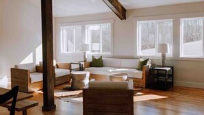 Een ruime woonkamer met een hoge plafond, grote ramen, een houten vloer en een gezellige zithoek met een bank en fauteuils.