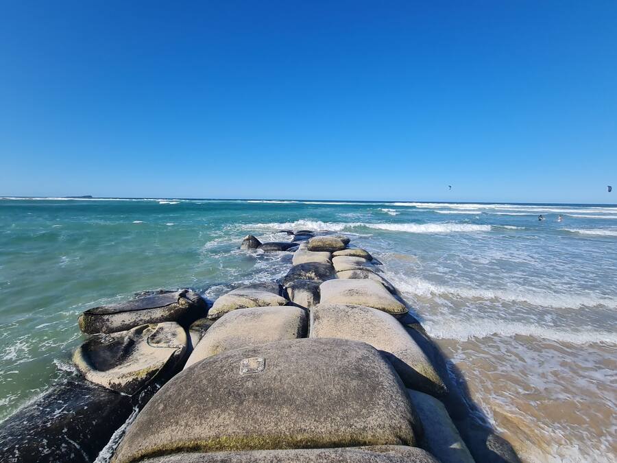 Un rompeolas de piedra que se extiende hacia una playa, con olas rompiendo contra él.