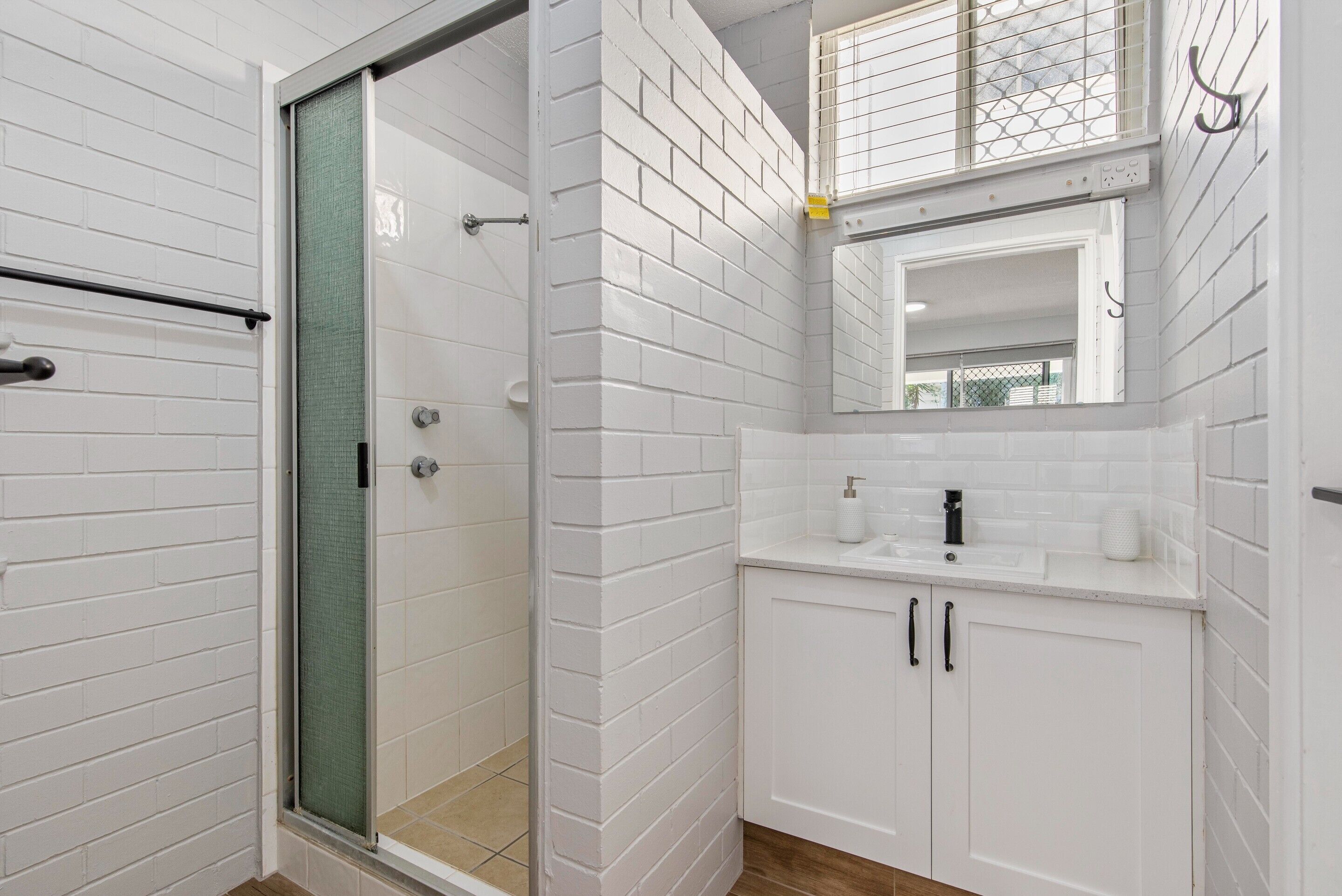 A bathroom with white tiled walls, a glass shower enclosure, a sink with a mirror above it, and wooden flooring.