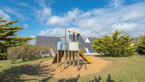 Un parque infantil con tobogán y estructuras para escalar, rodeado de árboles y un edificio al fondo.