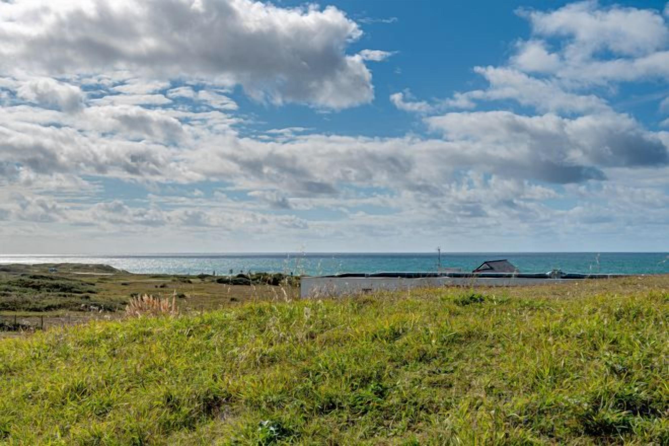 Un paysage côtier avec un premier plan herbeux, une étendue d’eau et une structure visible au loin.