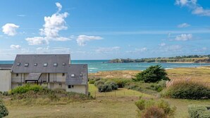 Un edificio costero con techo plano y varias ventanas, rodeado de terreno cubierto de hierba y vista al mar.