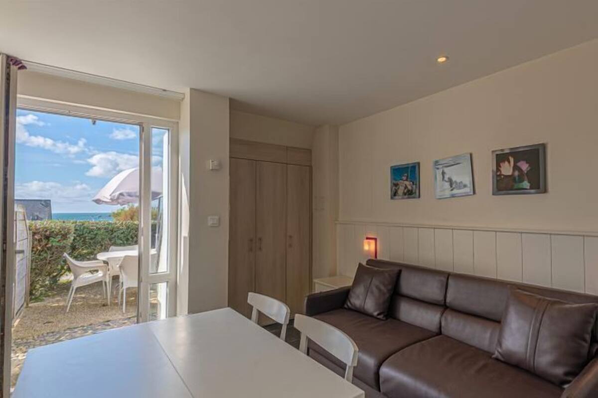 A room with a brown leather sofa, a white table, and a door leading to a balcony with a view of the sea.
