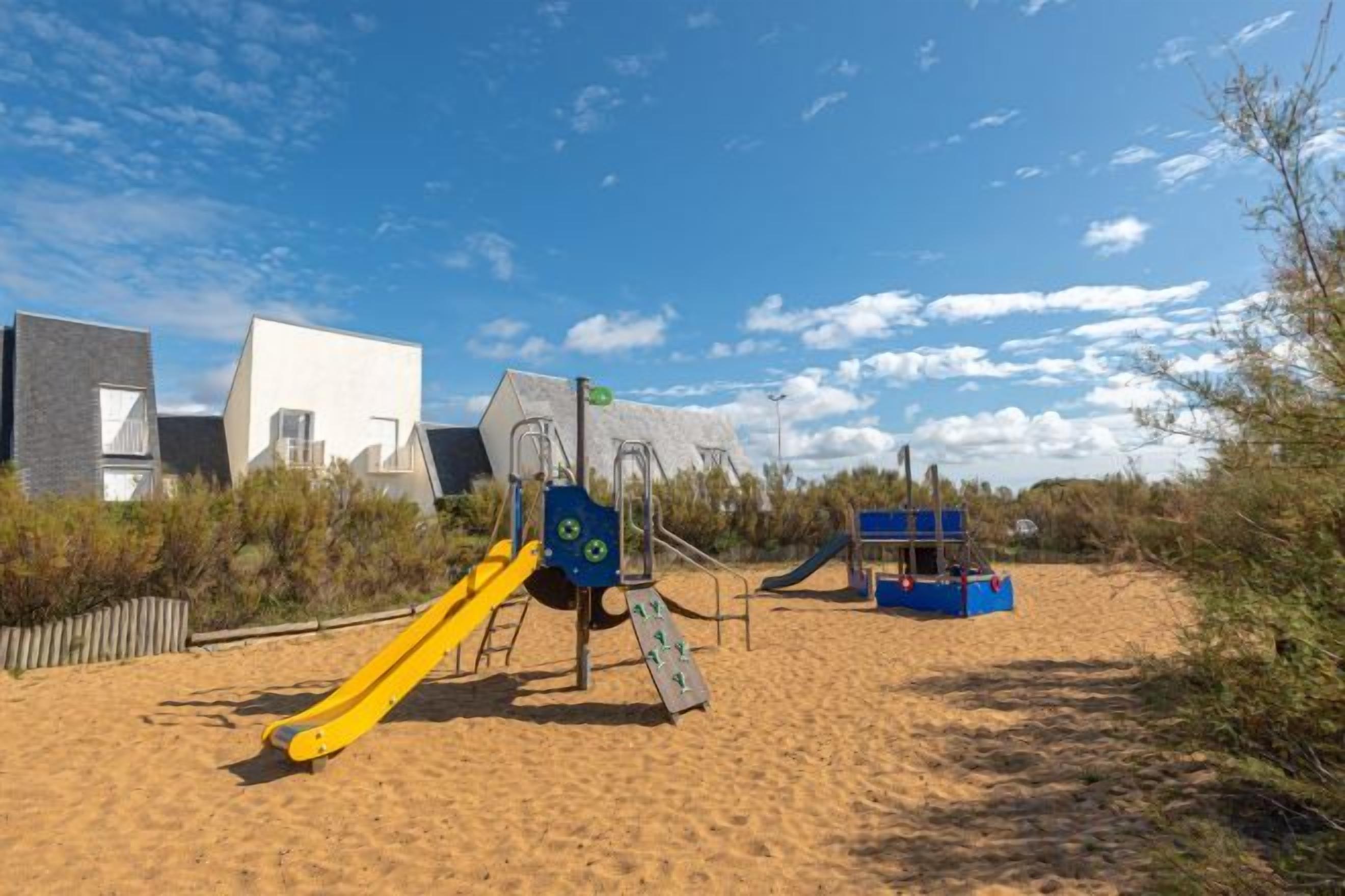 Une aire de jeux avec des toboggans et des structures d’escalade, située sur une plage de sable, avec des bâtiments en arrière-plan.