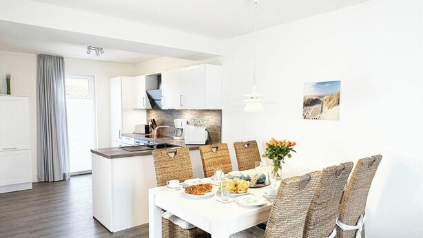 Une cuisine moderne avec des meubles blancs, un coin repas avec des chaises en osier et une vue sur la plage.