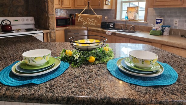 A kitchen with granite countertops, wooden cabinets, a stone backsplash, a hanging sign, and a window with curtains.