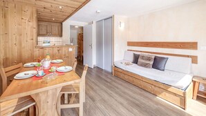 A modern, compact living space with a wooden dining table set for a meal, a sofa with a wooden headboard, and a kitchen area with wooden cabinets. - Cosy apartment for 4 people with WIFI, TV and balcony (Les Boisses)