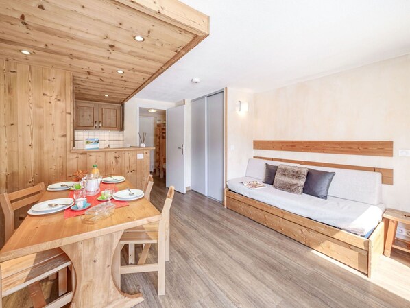 A modern, compact living space with a wooden dining table set for a meal, a sofa with a wooden headboard, and a kitchen area with wooden cabinets.