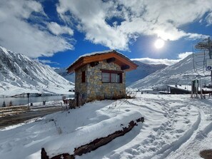 A stone cabin with a wooden roof, surrounded by snow-covered mountains and a clear sky.