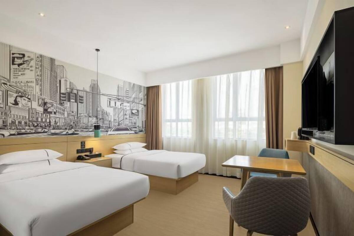 Habitación de hotel con dos camas, un escritorio, una silla, un televisor y un mural con vista a la ciudad.