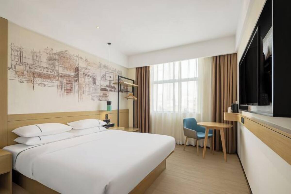 Habitación de hotel con una cama grande, un escritorio, una silla, un televisor y una pared con un mural de paisaje urbano.