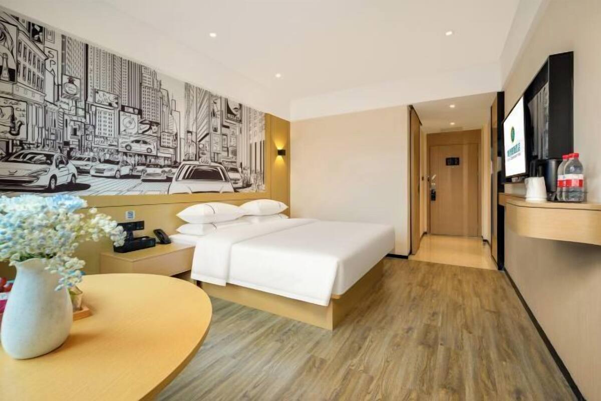 Habitación de hotel con cama, mesita de noche con un florero, televisor montado en la pared y un pasillo que conduce a otra habitación.