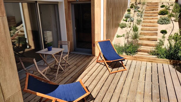 Une terrasse en bois avec deux chaises longues et une petite table, une porte-fenêtre coulissante et un jardin agrémenté de plantes et d’un escalier en bois.