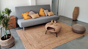 Un salon moderne avec un canapé gris, des coussins à motifs, un pouf tressé, un pouf rond, un grand vase et une plante en pot.