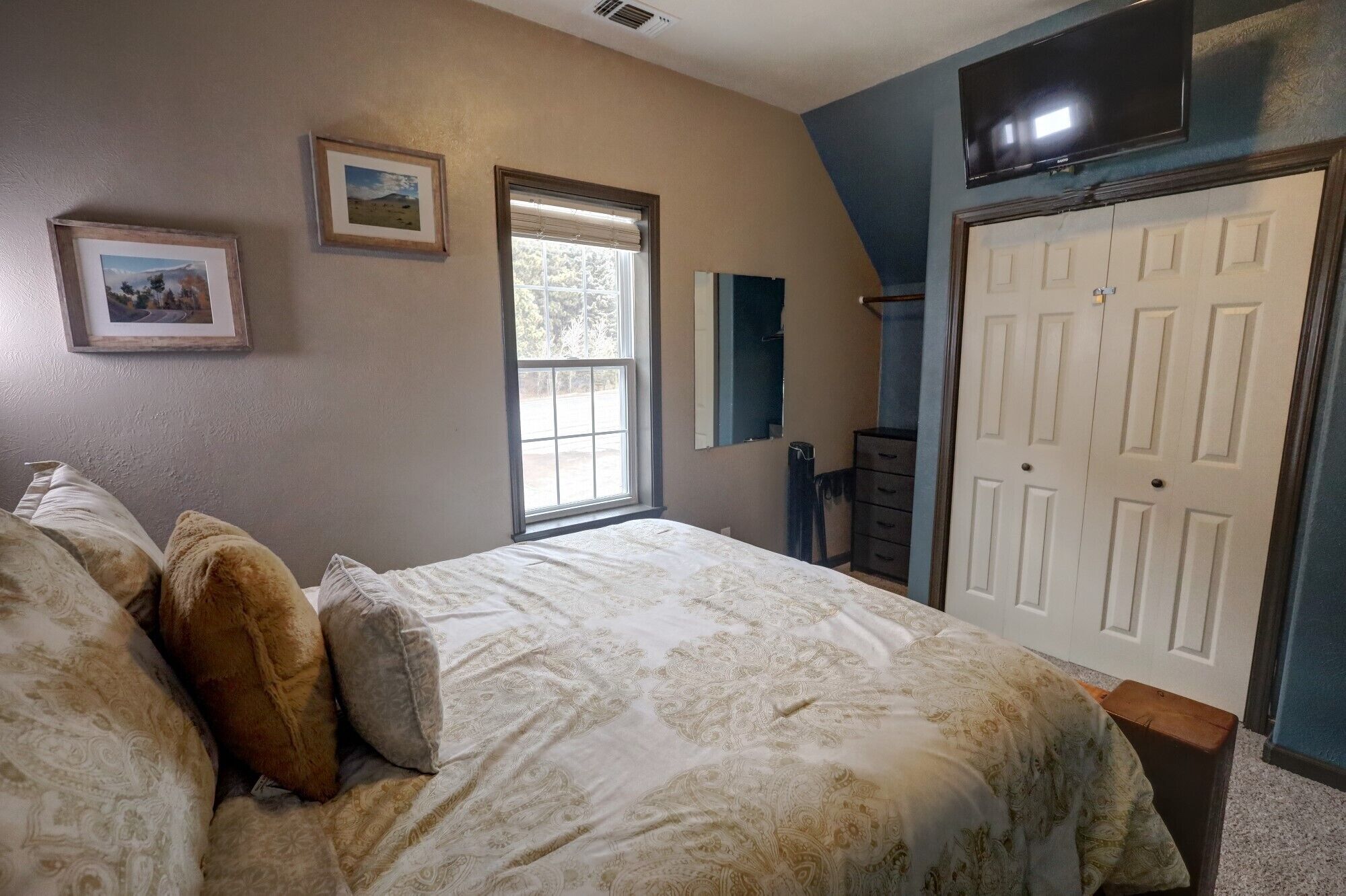 A bedroom with a bed, two framed pictures, a window, a mirror, and a door.