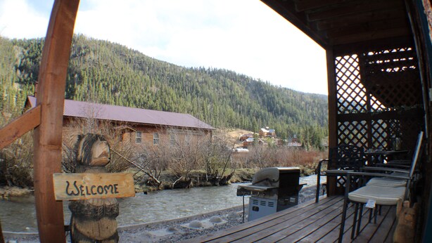 Une terrasse en bois avec une pancarte « Bienvenue », une rivière et une montagne boisée en arrière-plan.