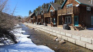 Une rangée de chalets en bois dotés de balcons, une rivière au premier plan et des arbres en arrière-plan.