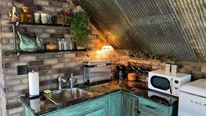 A kitchen with a brick wall, a wooden shelf with decorative items, a sink, a toaster, a coffee maker, and a refrigerator. - COUNTRY LACE Retro Place (Cabool)