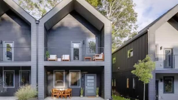 Una casa moderna de dos pisos con techo plano, ventanas amplias y un balcón con mobiliario exterior.