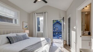Un dormitorio con una cama grande, un ventilador de techo, una ventana con persianas y vista a una casa azul en el exterior.