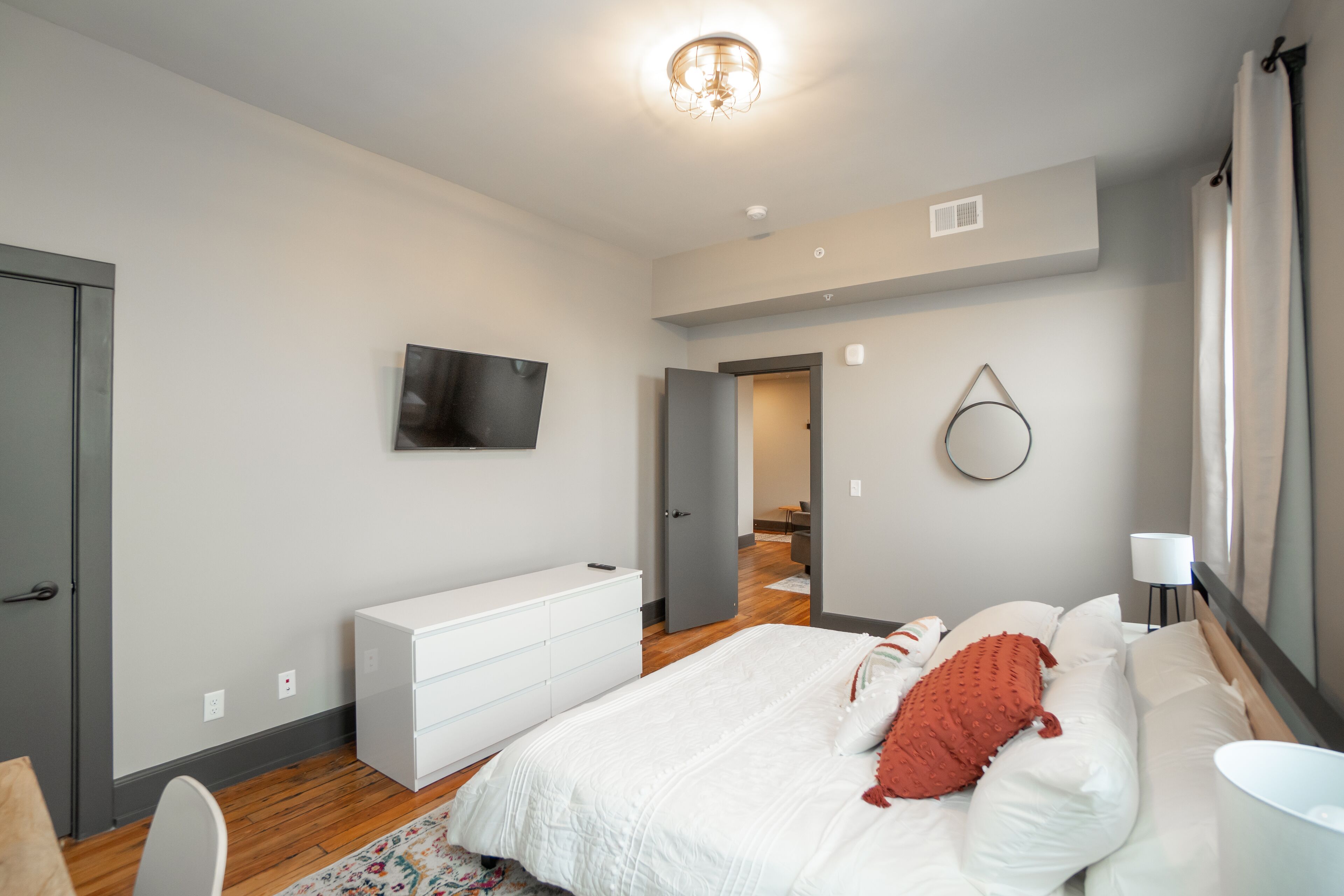 A modern bedroom with a large bed, a white dresser, a flat-screen TV, and a mirror on the wall.
