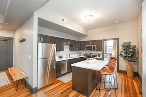 A modern kitchen with stainless steel appliances, a central island with bar stools, and a large window providing natural light. - Urban Lofts - Downtown Waterloo 2 Bedroom 3rd Floor (Waterloo)