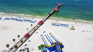 A beach with a long pier extending into the ocean, a lifeguard tower, and numerous beach umbrellas.