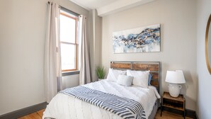 A bedroom with a bed, a nightstand, a lamp, a window with curtains, and a wall-mounted abstract painting.