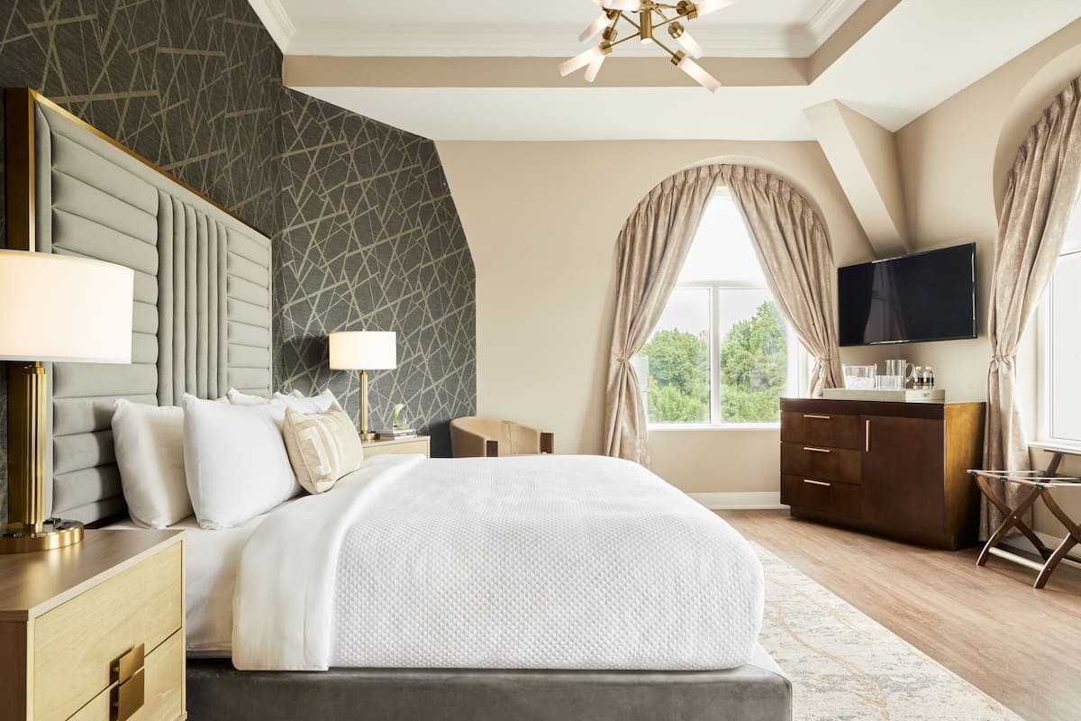 A modern hotel room with a large bed, a flat-screen TV, a desk, and a view of greenery outside.