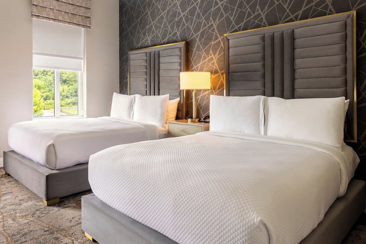 A hotel room with two beds, a nightstand with a lamp, and a window with a view of greenery.