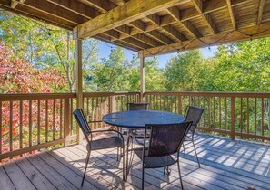 Une terrasse extérieure couverte, dotée d’une table ronde et de quatre chaises, entourée d’une balustrade en bois et d’arbres.