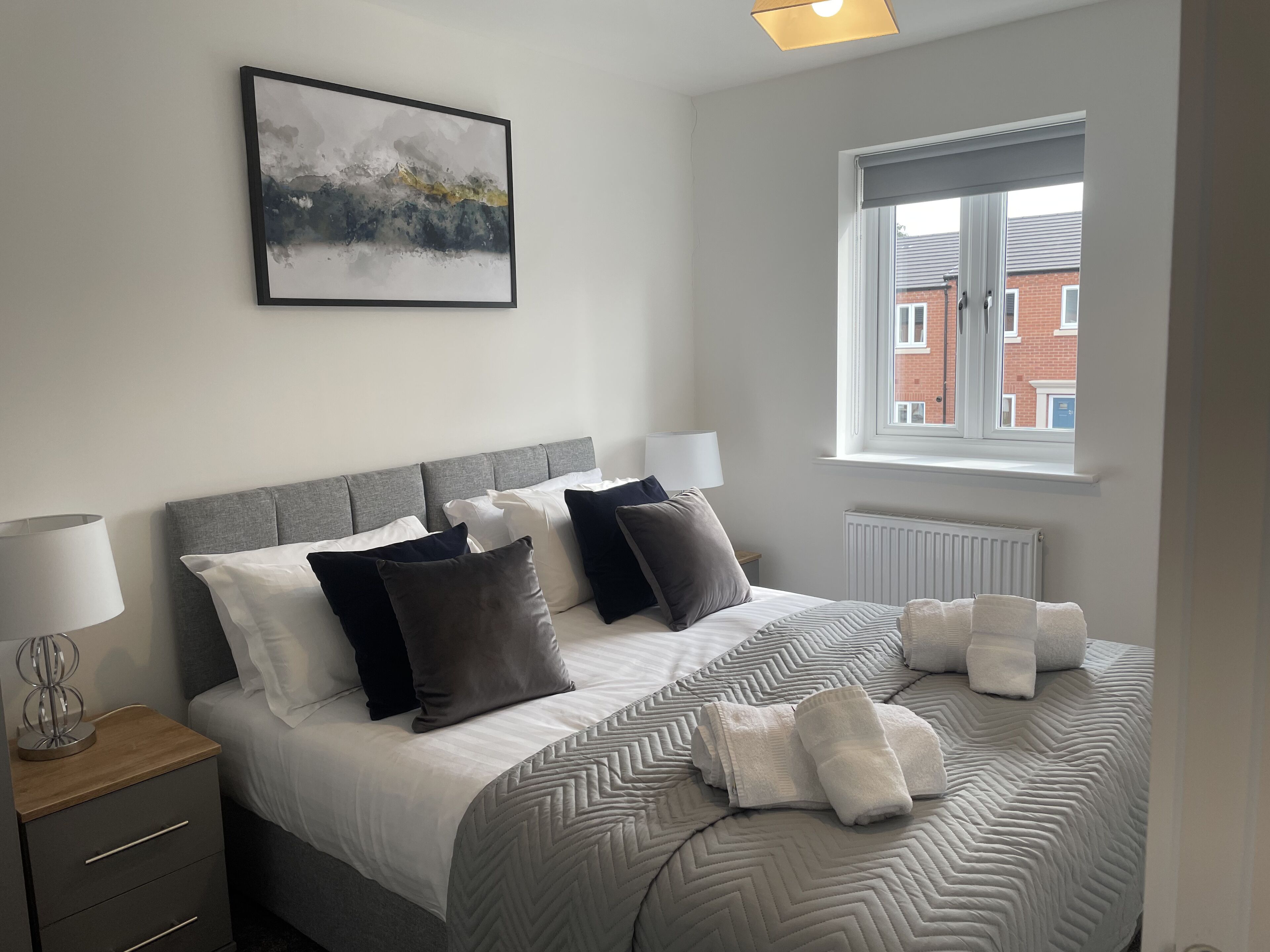 A neatly made bed with a grey headboard, white and dark grey pillows, and a grey patterned bedspread. There is a window with a view of buildings outside.