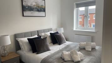 A neatly made bed with a grey headboard, white and dark grey pillows, and a grey patterned bedspread. There is a window with a view of buildings outside.