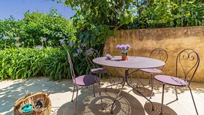 A round metal table with four chairs, a vase with purple flowers, and a wicker basket with a blue object on a patio with a stone wall and greenery in the background.